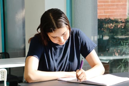 A student sits at a table indoors, focused on writing in a notebook with a pen as natural light comes through nearby windows.