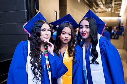 Three students stand together wearing blue graduation caps and gowns, smiling and posing backstage before a graduation ceremony.