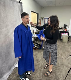 student in a gown taking his graduation picture while a teacher helps him