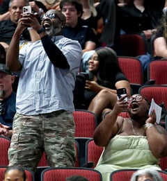 two adults in the arena stands cheering and taking photos of their graduate