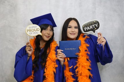 two students in blue graduation cap and gowns wearing orange feathers and holding signs (1)